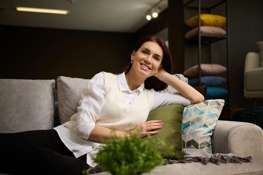 Beautiful woman sitting on a new sofa on sale, examining it before buying. Attractive woman shopping for upholstered furniture at shopping mall. Consumerism, interior design and new home concept