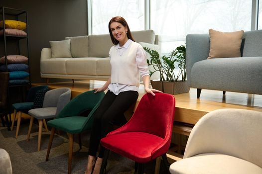 Charming sales assistant in casual clothes, standing near velour stylish chairs in the furniture shopping mall, smiles looking at camera while presenting the new collection of upholstered furniture