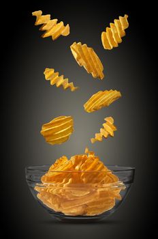 Tasty, grooved potato crisps are falling down in a glass bowl against a black background with copy space for your text or images. Crispy, palatable chips. Shadow. Advertising concept. Close-up shot.