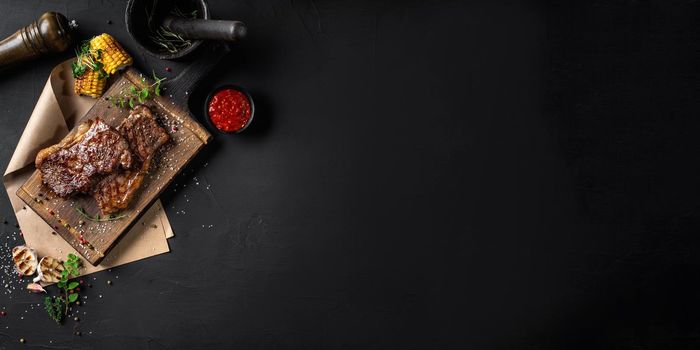 Fried sappy steaks with herbs and spices on a wooden board, parchment, grilled garlic and corn, sprouts, spice chopper, sauce in a small dark bowl, mortar with pestle on a black background. Cooking, restaurant concept. Close-up shot. Top view. Dry aged