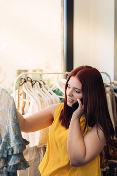 Young smiling and happy red-haired business woman buying clothes in a fashion shop. talking on mobile phone. shopping concept. Natural light, sun rays, display with clothes, clothes rack, customers, clothes, vertical view. dressed in yellow t-shirt.