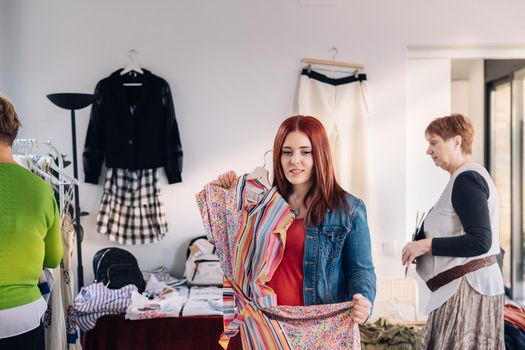 young woman trying on a dress in front of a mirror. red-haired girl shopping in a fashion shop. shopping concept. leisure concept. Natural light, sunbeams, display, clothes rack, clothes, vertical view, space to copy. customer in blue denim jacket, red t-shirt. out of focus motion effect on background.