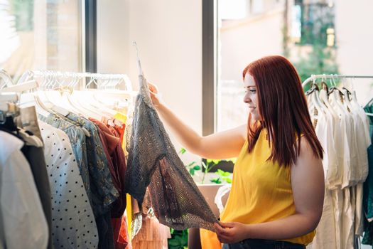 Young smiling and happy red-haired girl looking at clothes on a hanger to buy, in a fashion shop. concept of shopping. Natural light, sun rays, display with clothes, clothes rack, clothes, vertical view. dressed in yellow t-shirt.