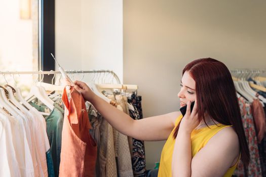 Young red-haired business woman buying clothes in a fashion shop. talking on mobile phone. shopping concept. Natural light, sun rays, display with clothes, clothes rack, customers in background walking, clothes, vertical view. dressed in yellow t-shirt.