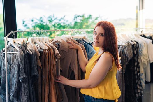 Young smiling and happy red-haired woman, happy and enjoying a day of shopping, in a fashion shop. looking at camera. concept of shopping. concept.natural light, sun rays, display with clothes, clothes rack, shop window, clothes, Horizontal.