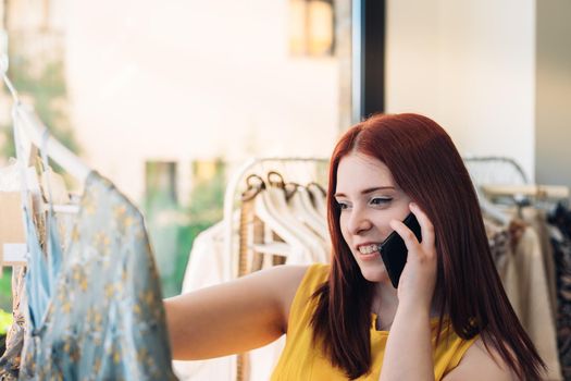 Young red-haired business woman buying clothes in a fashion shop. talking on mobile phone. shopping concept. Natural light, sun rays, display with clothes, clothes rack, customers in background walking, clothes, vertical view. dressed in yellow t-shirt.