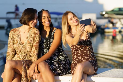 Positive young multiracial female friends in dresses taking self portrait while sitting together near river with boats on summer day