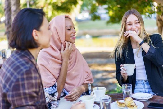 Cheerful young multiracial diverse ladies in stylish clothes laughing and chatting while having coffee break in outdoor cafe on sunny day