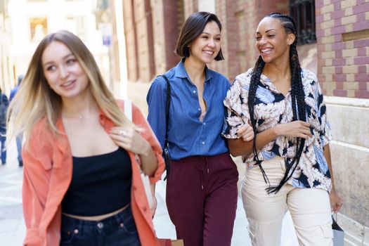 Group of smiling diverse female friends with shopping bags strolling on street near building during shopping in city on summer day