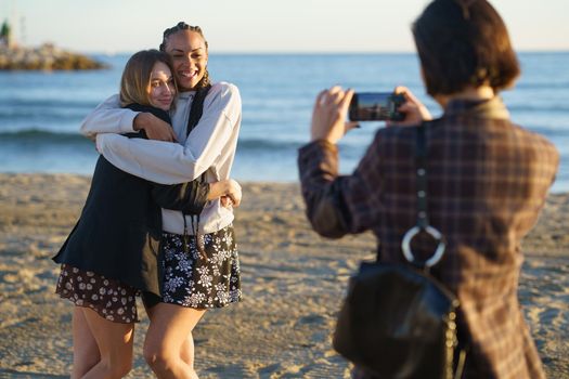 Anonymous female with smartphone taking picture of smiling multiethnic girlfriends hugging on sandy coast near waving sea on summer day