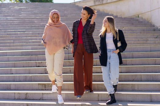 Confident young happy multiracial women students in casual outfits and hijab smiling while walking downstairs in university campus on sunny day