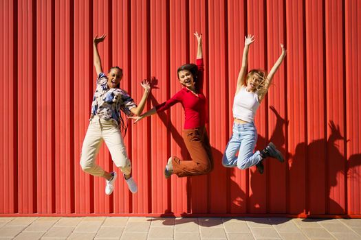 Full body of joyful diverse women looking at camera and jumping high above ground near red wall while having fun on sunny street