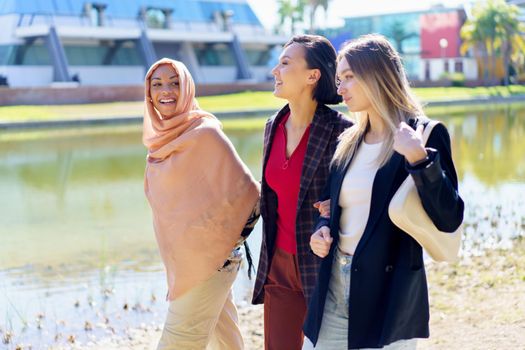 Cheerful young multiracial women friends in stylish clothes smiling and chatting while walking together on embankment near river on sunny day in city