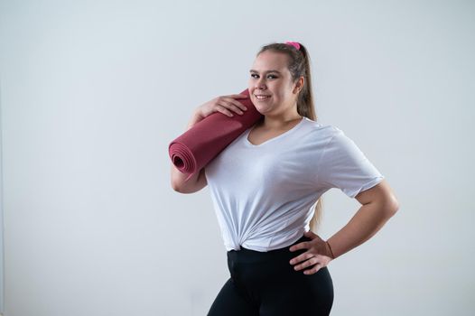 Young fat caucasian woman holding a sport mat. Charming plus size model in sportswear stands on a white background.