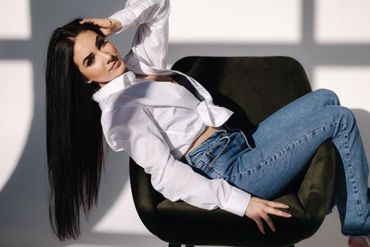 Female sitting on green chair. Her black hair is hanging down. Pretty woman in denim and white shirt.