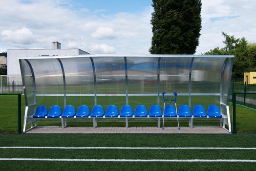 Crutch at blue plastic seats on outdoor stadium players bench, chairs with new paint below transparent plastic roof. 