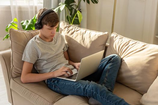 Portrait of teenager boy in a gray t-shirt and blue jeans, lying on a sofa in a room with plant, wearing black headphones on his head, looks into a gray laptop, on his knees. Copy space