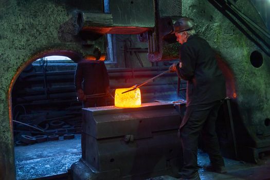 gray haired blacksmith worker taking large piece of hot steel with big forceps in old factory in Tula, Russia - February 1, 2020