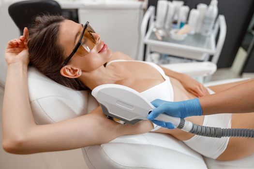 Female client in eyeglasses during armpit photo epilation procedure with ipl machine in beauty salon.