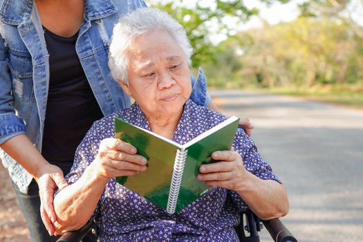 Asian senior or elderly old lady woman patient reading a book while sitting on wheelchair in park : healthy strong medical concept