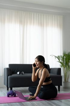 Portrait asian sporty woman in sportswear working out and using laptop and call phone at home in living room, sitting on the floor with dumbbells on yoga mat. Sport and online training concept.