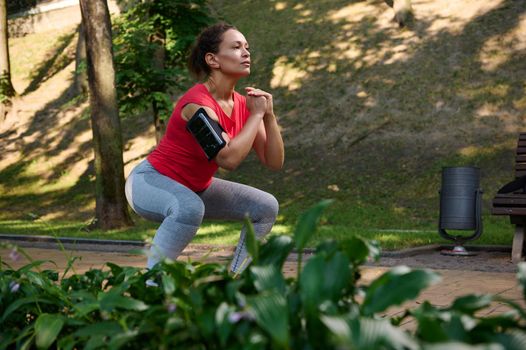 Determined athlete, sportswoman exercising outdoor, doing deep squats in the city park. Enjoy summer days and keep your body fit. Sport, fitness, functional body weight training concept