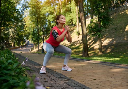Beautiful confident middle-aged fit woman performing deep squats in the city park, enjoying outdoor workout on a beautiful sunny summer day. Sport, fitness, functional body weight training concept