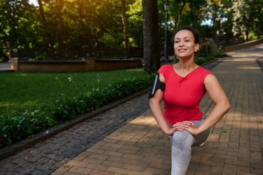 Beautiful middle aged sporting woman exercising outdoor, doing lunges, stretching her leg muscles while exercising in the city park on warm sunny summer day