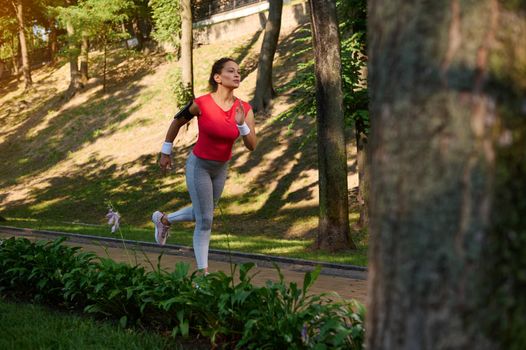 Full length. Active middle aged Caucasian woman, sportswoman, jogger exercising outdoors, running on a treadmill in a city park on a sunny warm summer day. Sport, fitness, functional training concept