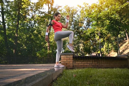 Full length portrait of a charming active Hispanic woman in tight sportswear exercising outdoors doing leg muscle exercises on the steps of a forest park on a summer day. Keep your body fit