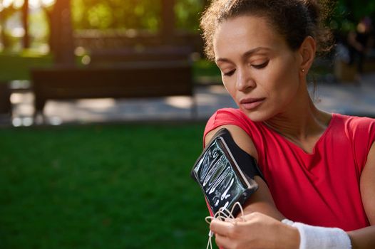 Close-up portrait of a young athletic woman, a runner in a bright red t-shirt, connects headphones to a mobile phone and sets up the application before jogging in the park on a sunny summer day