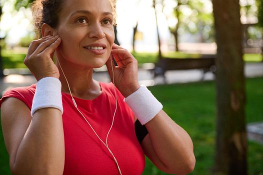 Attractive middle aged active woman, sportswoman putting on earphones, wearing bright red t-shirt and white terry wristbands, getting ready for outdoor workout and morning run