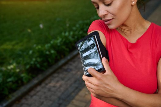 Close-up of an attractive woman, athlete adjusting the mobile app in her mobile phone in the smartphone holder, standing in the city park, getting ready for morning jog