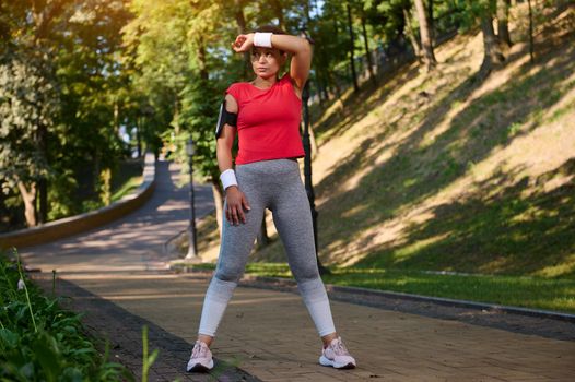 Full length portrait athlete woman, runner wiping the sweat from her face using terry wristband while running in city park, keeping her body fit. Sport, fitness, healthy and active lifestyle concept