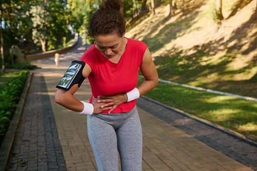 Determined middle aged fit woman, runner, sportswoman holds her stomach, has side cramps, pain during running training . Sports injuries concept