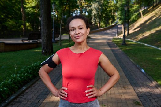 Charming muscular woman with healthy aesthetic body and glowing skin, athlete in red t-shirt and smartphone holder posing with arms on waist looking at camera standing on the city park treadmill
