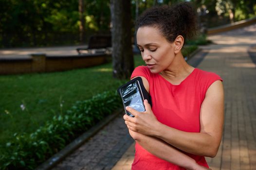 Attractive stunning fit woman in tight red t-shirt, athlete adjusting the mobile app in her mobile phone in the smartphone holder, standing in the city park, getting ready for morning jog