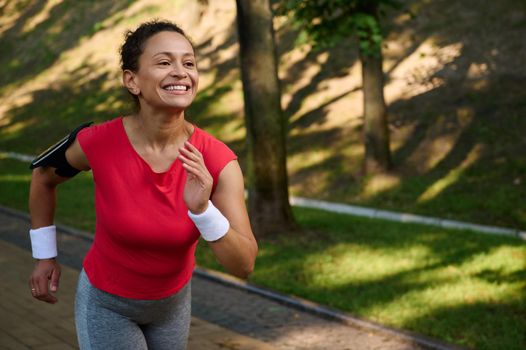 Close-up young African American fit woman, athlete in tight sportswear running along the treadmill of a city park, enjoying cardio workout on a beautiful summer sunny day