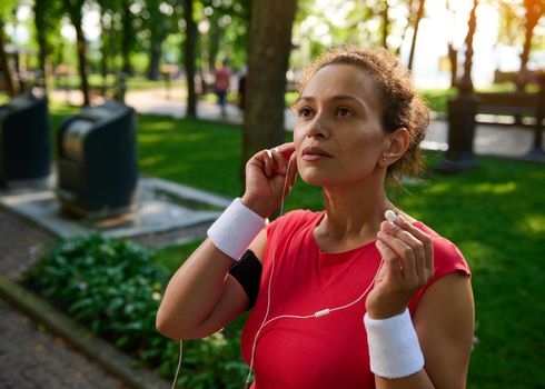 Active beautiful African American sportswoman, runner, athlete wearing white terry wristbands, putting on earphones, listening to the music while jogging along the city park early in the morning