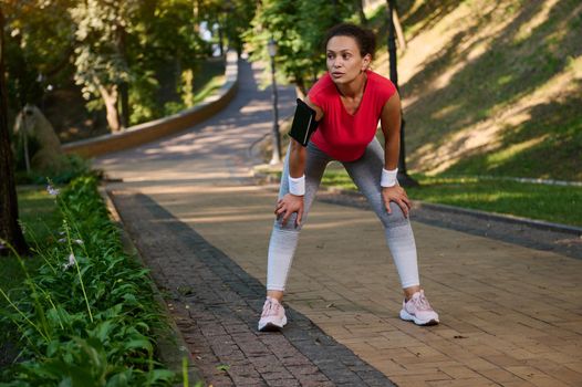 Female runner, athlete, sportswoman resting in the city park while feeling exhausted during jogging on the treadmill on a warm sunny summer day. Body weight training, slimming and dieting concept