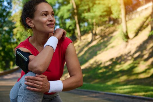 Close-up portrait of beautiful Hispanic woman, athlete in sportswear relaxing during jog in the city park, smiles looking aside, enjoying morning workout on summer sunny day