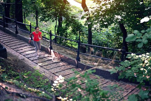 Overhead view of a middle-aged sportswoman running on stairs along railing in forest park. Cardio workout, jog, active and healthy lifestyle concept