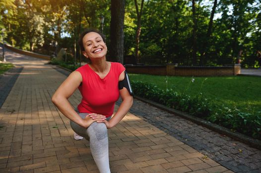 Happy cheerful African woman doing lunges while exercising outdoor, smiles cutely with beautiful toothy smile looking at camera, enjoying her active and healthy lifestyle