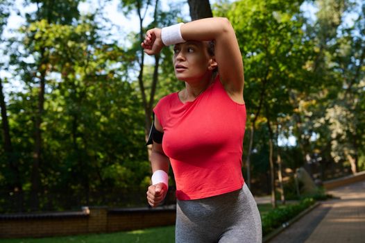 Close-up. Beautiful athlete woman, runner wiping the sweat from her face using terry wristband while running in city park, keeping her body fit. Sport, fitness, healthy and active lifestyle concept