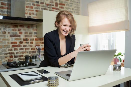 A woman works remotely on a laptop. A laughing girl with braces holds a cup of coffee listening to the report of a colleague at an online briefing at home. Lady preparing for a lecture on video call.