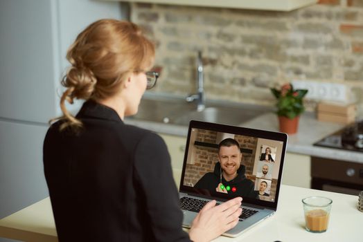 Back view of a businesswoman who speaks with colleagues during a video conference on a laptop. A female boss talks on a video call with her employees engaged in an online business briefing at home.
