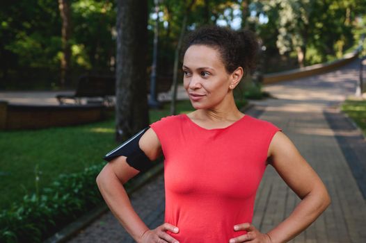Charming muscular woman with healthy aesthetic body and glowing skin, sportswoman in red t-shirt and smartphone holder, posing with hands on waist looking away, standing on treadmill of a city park