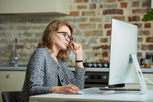 A pretty woman in glasses works remotely on a desktop computer in her studio. A female boss searching for information at a video conference at home. A female professor preparing for an online lecture..