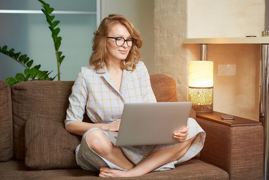 A young woman in a dress sitting on the couch with legs crossed works remotely on a laptop in her apartment. A girl during a webinar at home. A female student in glasses listening to an online lecture