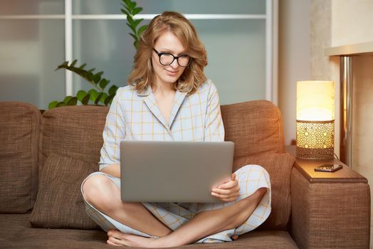 A young woman in glasses works remotely on a laptop in her apartment. A cute girl during a video conference with her partners at home. A female student with a smile listening to an online lecture.